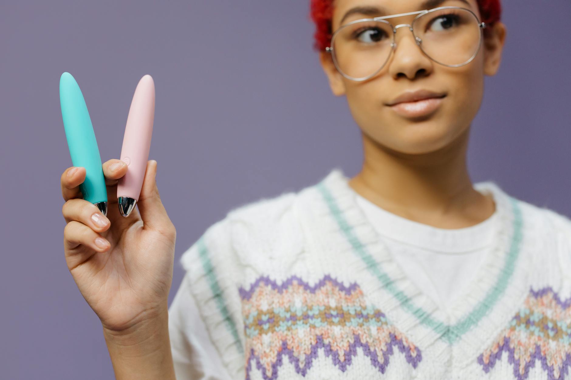 A woman in eyeglasses holding two colorful vibrators against a purple background, representing sexual choice and self-pleasure
